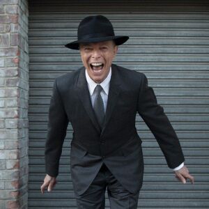 An image of David Bowie in a stylish dark suit and hat. He is leaning towards the camera and seems to be laughing riotously towards it.